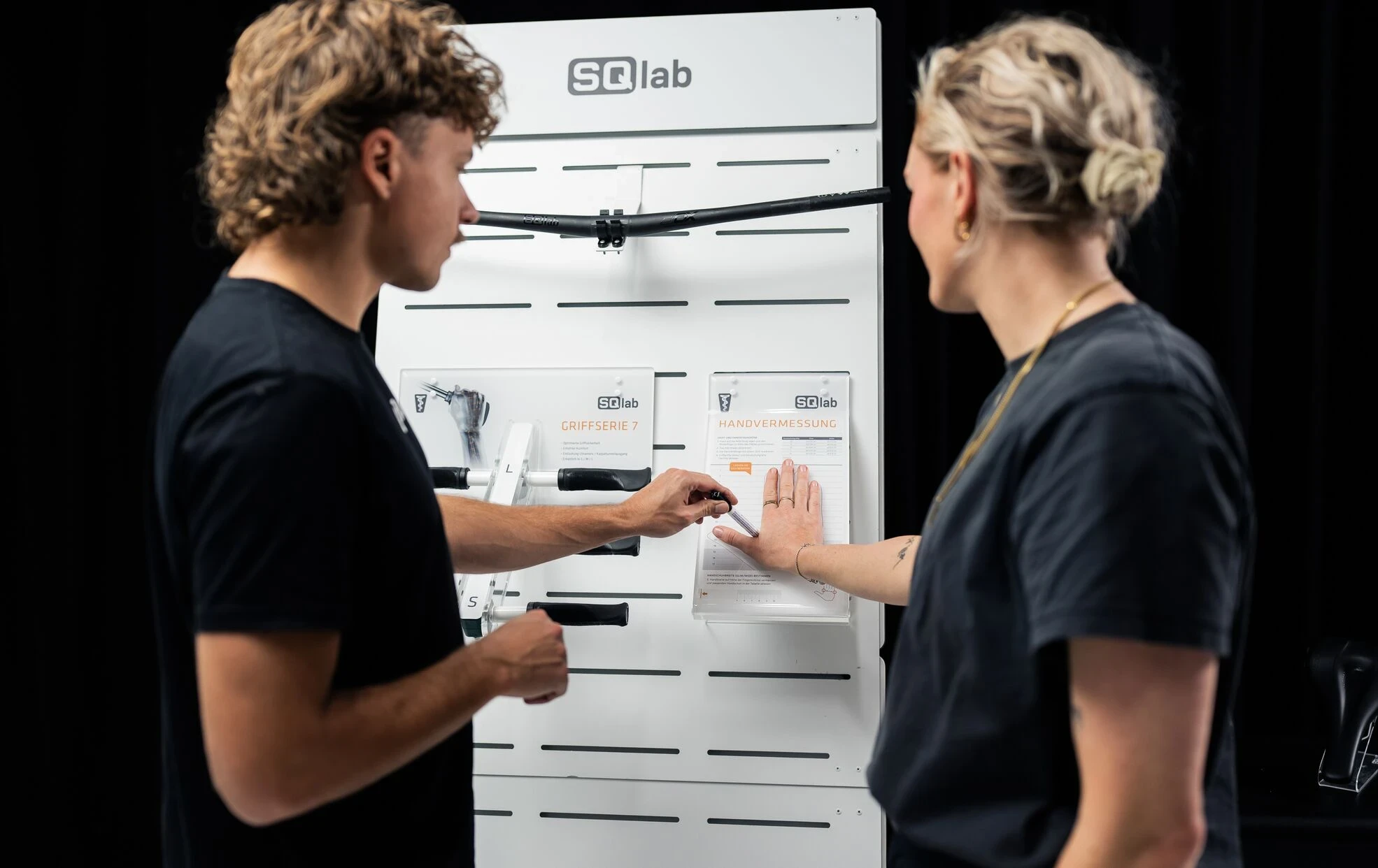 Zwei Personen in schwarzen T-Shirts beraten sich vor einer wei&szlig;en SQlab Produktwand.